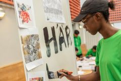 Under ledning av Anders ”Bleckan” Bleckhorns från Salong Betong kommer nio ungdomar att gestalta budskapet i en 25 meter lång målning på planket runt bygget av det nya polishuset i Rinkeby. Här jobbar Fatou Njie med en skiss till konstverket. Foto: Niklas Palmklint