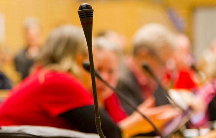 I förgrunden en mikrofon, i disig bakgrund skymtar mötesdeltagare