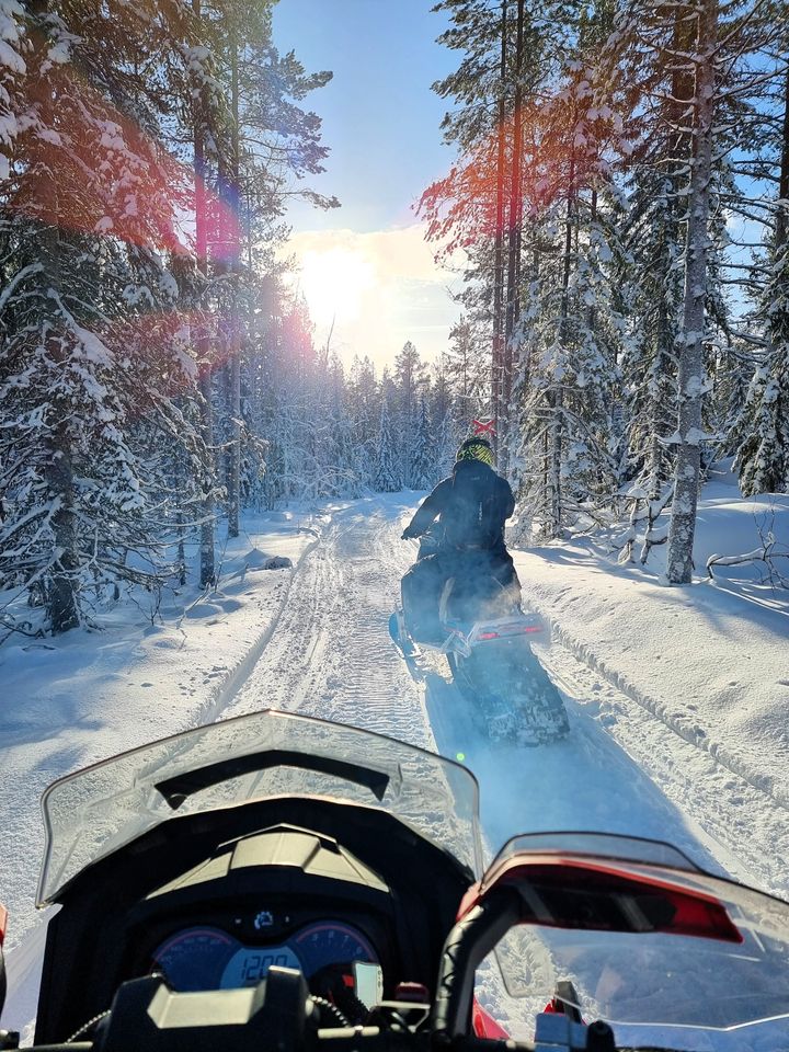 Två snöskotrar efter en fin skoterled, solen skiner och vid leden syns snötäckta granar.