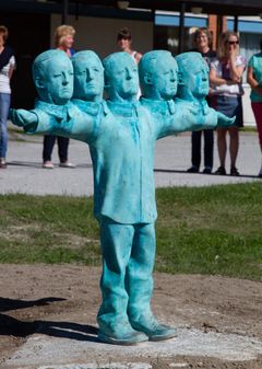 Grön skulptur företällande en pojke med fem huvuden. De extra huvudena är placerade, 2 vardera, på pojkens utsträckta armar.