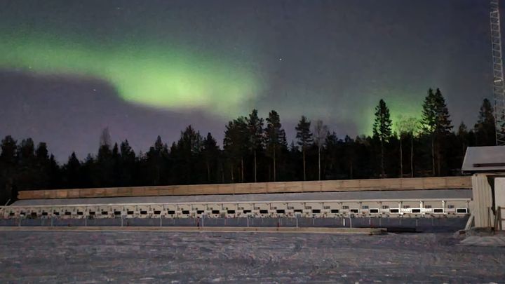 Norrsken lyser upp himlen över en skidskyttevall omgiven av träd på kvällen.