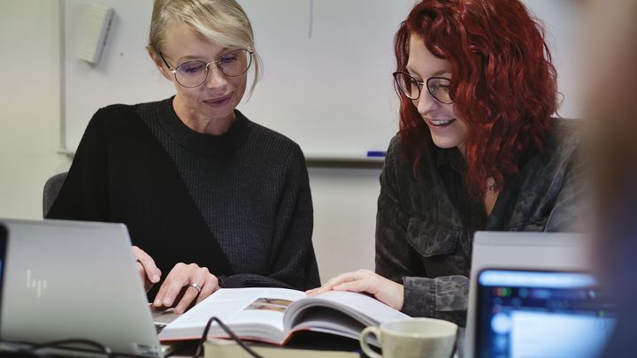 Två kvinnor studerar vid dator och uppslagan böcker.