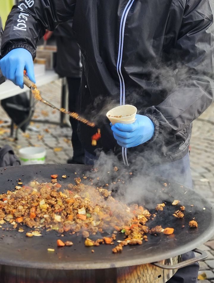 En person lagar mat på en stor stekhäll utomhus, iklädd svarta kläder och blåa handskar. Rök stiger från maten.