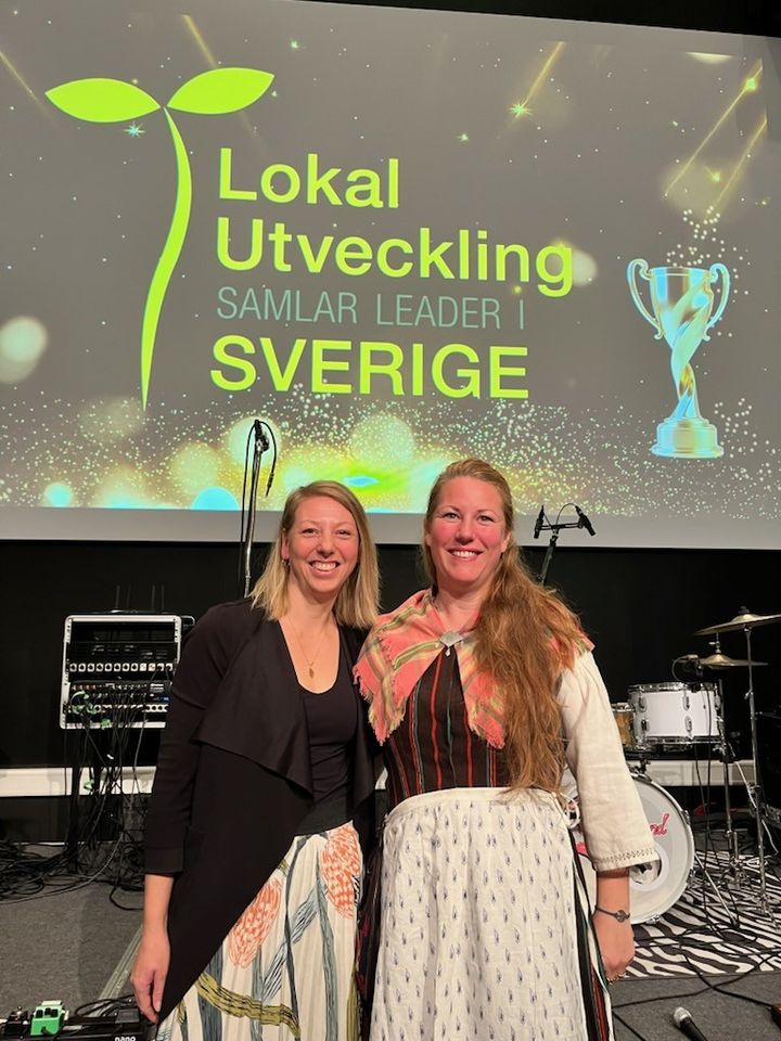 Två kvinnor står på en scen framför en stor skärm med texten ”Lokal Utveckling samlar leder i Sverige”.