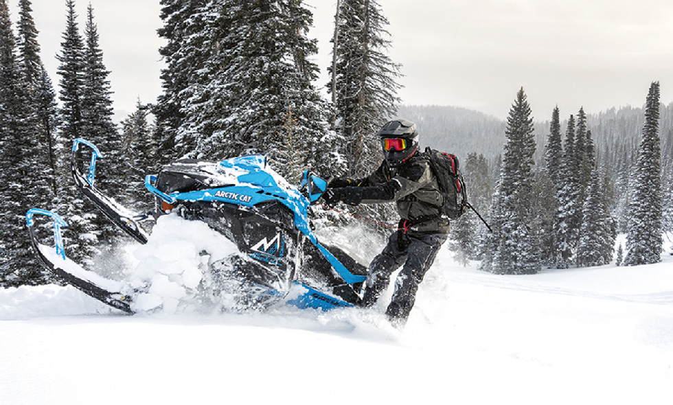 Skoteråkare kör en blå och svart Arctic Cat i djup snö bland snötäckta granar.