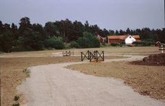 Nyanlagd grusväg med små träbroar över öppna diken, omgiven av oplanterad mark och skog i bakgrunden.