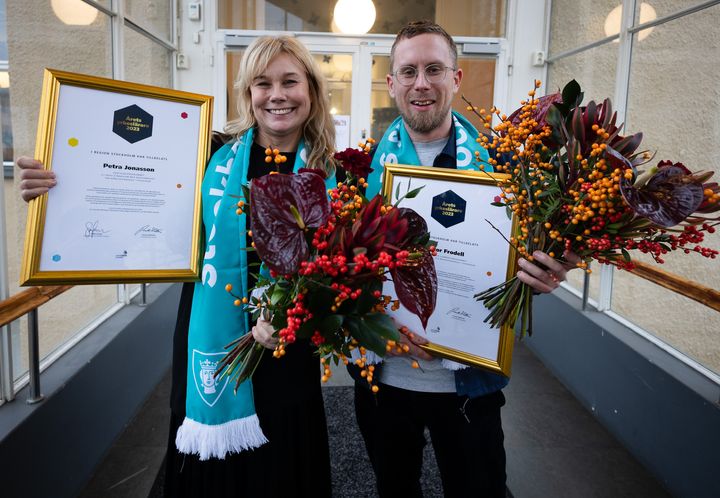 Två glada lärare visar upp diplom och blommor framför entre till S:t Eriks gymnasieskola