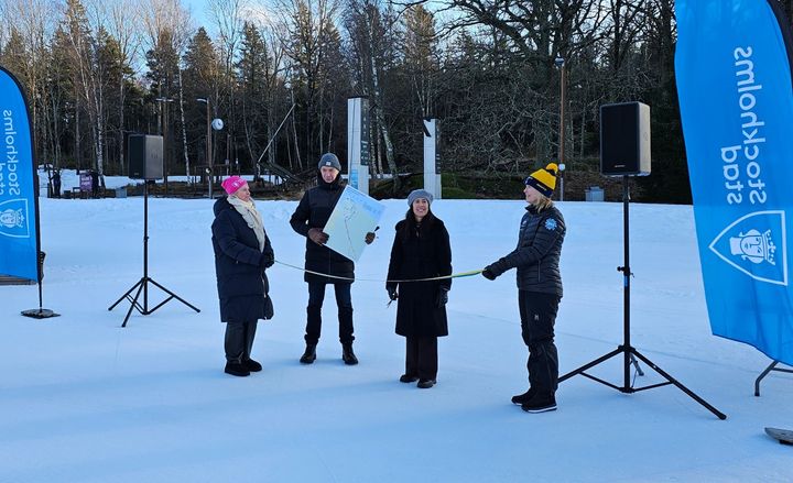 Fyra personer på snötäckt yta, två håller i ett blågult band som en tredje person ska klippa av