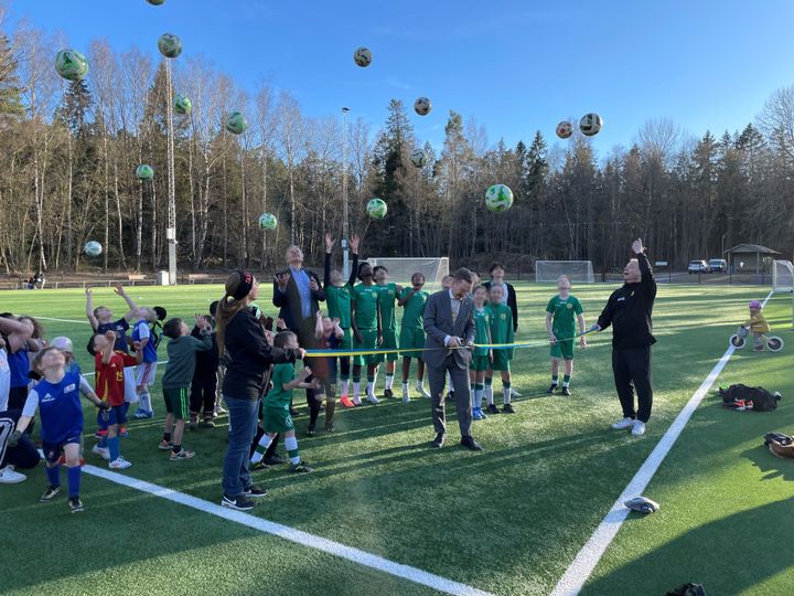 Barn som står på en fotbollsplan och kastar upp massa bollar, samtidigt som ett blågult band klipps av.