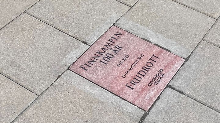 En rosa-röd trottoarsten med text om 100-årsjbilerande Finnkampen, på en trottoar med gråa trottoarstenar.