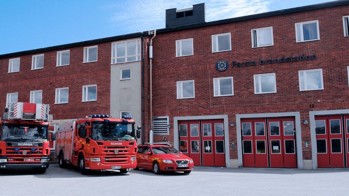 Farsta brandstation är en tegelbyggnad med stora röda portar till vagnhallen. Framför brandstationen står tre olika typer av bilar som räddningstjänster använder vid larm.