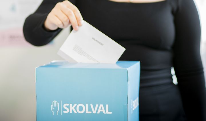 Person som lägger i ett valkuvert i ljusblå papplåda med texten skolval.