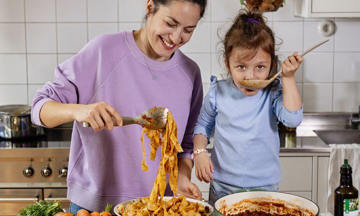 En vuxen tjej och en flicka i ett kök. De lagar pasta tillsammans och flickan provsmakar.