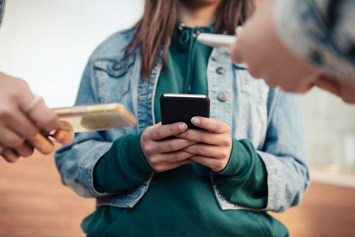 Ung person står och håller i en mobiltelefon. Fler unga, också med mobil, står runt omkring.