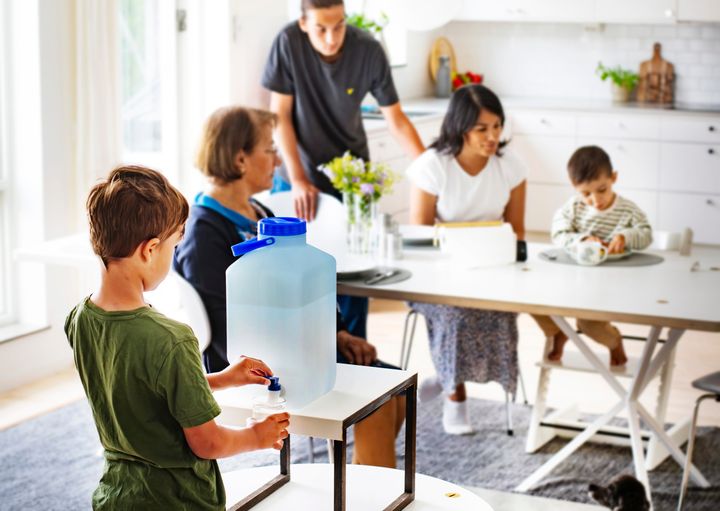 Familj samlad runt köksbord. Pojke fyller ett glas från en dunk med vatten.