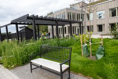 En modern byggnad med stora fönster, en pergola och en svart bänk i en grön trädgård.