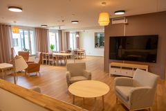 Ett modernt och ljust sällskapsrum med bekväma fåtöljer, ett runt bord, en stor TV och matbord i bakgrunden.