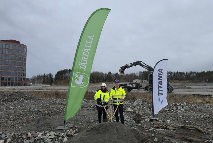 Två personer med hjälmar håller i en spade på en byggarbetsplats, omgivna av flaggor för Järfälla och Titania.