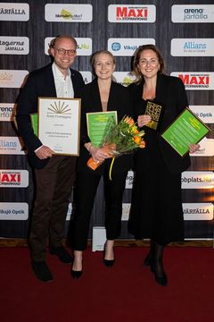 Tre personer står på en röd matta och håller diplom och blommor framför en fotobakgrund med olika logotyper.