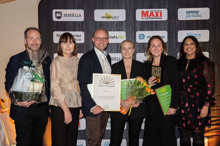 Sex personer poserar leende med diplom och blommor vid en prisutdelning.
