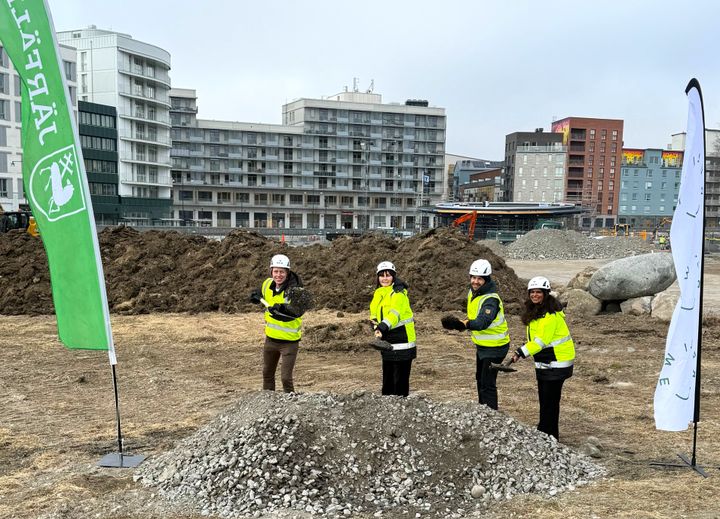 Fyra personer i varselkläder och hjälmar står vid en byggarbetsplats med spadar.