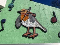 Mosaik av en fågel som spelar fiol mot en grön bakgrund, med musiksymboler runt omkring.