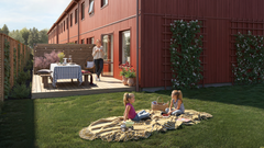 Två barn har picknick på en gräsmatta framför ett rött radhus. En person står på altanen.