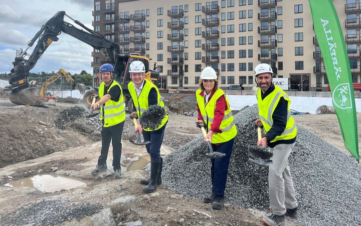Fyra personer deltar i en symbolisk byggstartsceremoni med skyfflar, iklädda hjälmar och reflexvästar. En grävmaskin och ett byggprojekt syns i bakgrunden.