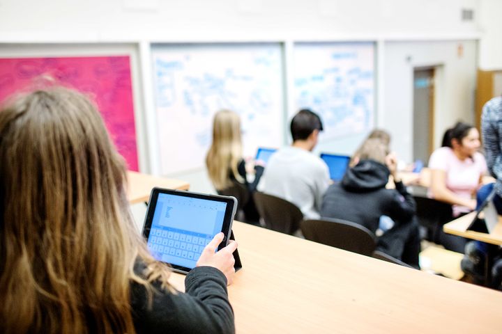 En elev med en surfplatta i ett klassrum, fler elever i bakgrunden.