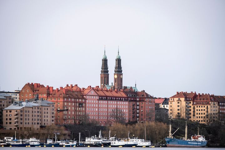 Stockholmsilhuett med två kyrktorn som reser sig över byggnaderna. Flera båtar syns i förgrunden.