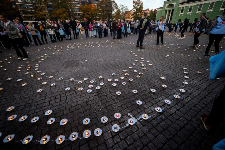 A large gathering of people stands around concentric circles of lit candles on a cobblestone pavement during a vigil.