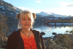 A person standing outdoors near a lake with mountains in the background.