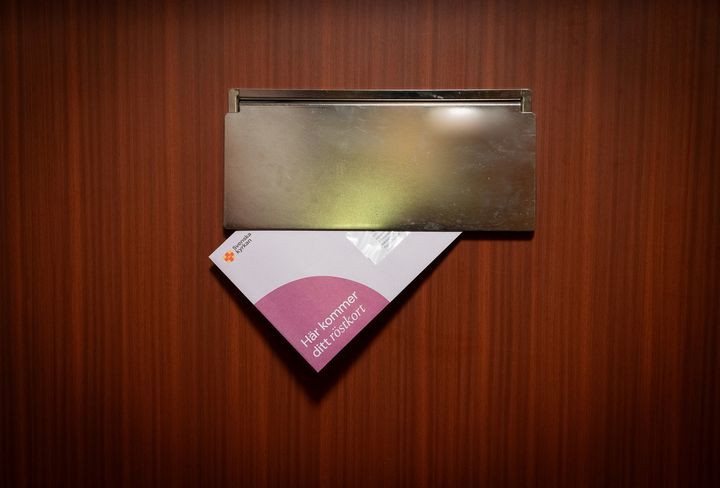 Envelope partially inserted through a brass mail slot in a wooden door.