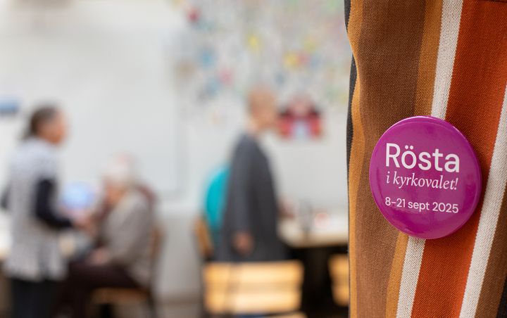 A pink button with the text "Rösta i kyrkovalet! 8-21 sept 2025" is pinned to a striped fabric. Blurred people in the background.