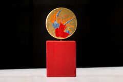 En statyett med ett runt, färgglatt konstverk föreställande en ängel, placerad ovanpå ett rött, rektangulärt fundament.