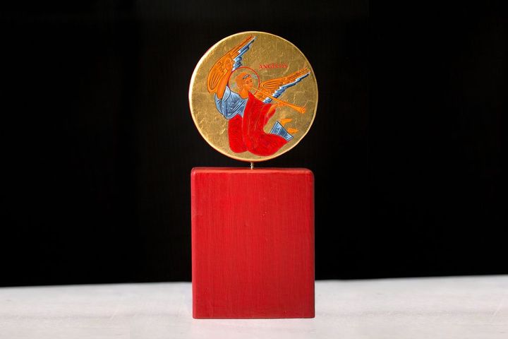 En statyett med ett runt, färgglatt konstverk föreställande en ängel, placerad ovanpå ett rött, rektangulärt fundament.