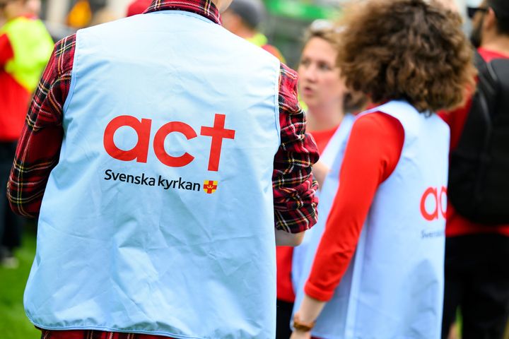 Personer i västar med texten "Act Svenska kyrkan" samlade utomhus.