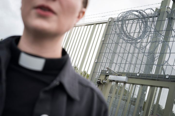 A person wearing a clerical collar stands in front of a tall metal gate topped with barbed wire.