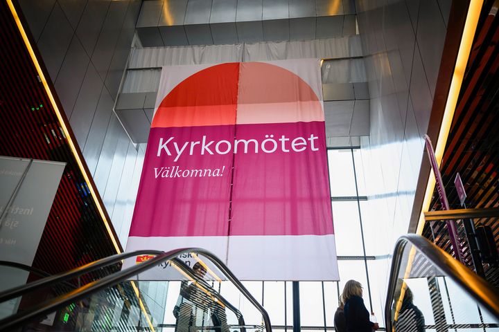 En stor banderoll med texten "Kyrkomötet Välkomna!" hänger i en modern byggnad.