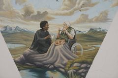 Alt text: En målning föreställande en präst och en äldre person i rullstol som dricker kaffe tillsammans i en naturskön miljö.