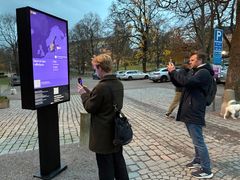 En digital skärm utomhus visar en lila karta över Sverige med statistik om tända ljus. Två personer fotograferar skärmen med sina mobiltelefoner på en stenlagd gata, med parkerade bilar och träd i bakgrunden.