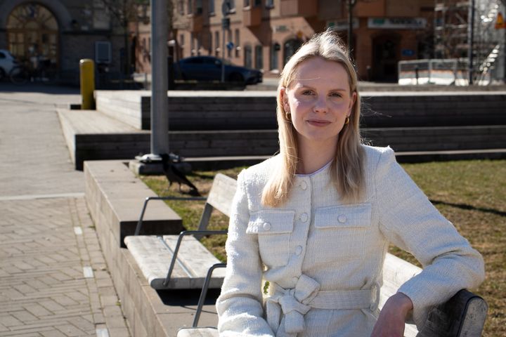 Kvinna i vårsolen på en parkbänk. Hon har ljust långt hår och tittar leende in i kameran.