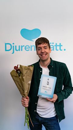 En man står framför en vägg med texten "Djurens Rätt" och håller en blombukett och ett diplom.