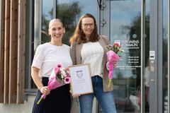 Två kvinnor står framför en byggnad, hållandes blommor och ett diplom.