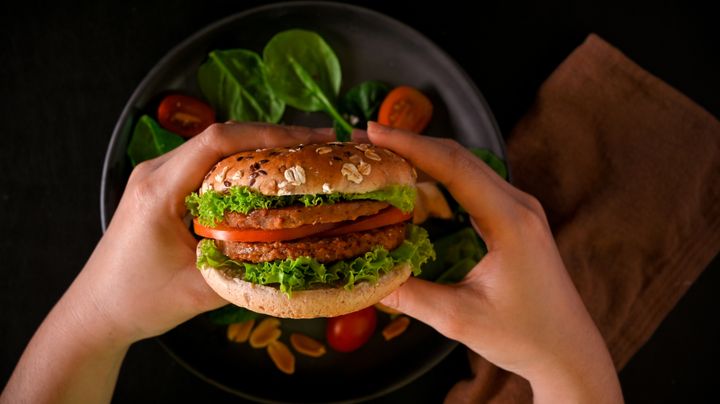 Två händer håller en hamburgare med sallad och tomat, på en tallrik med spenat och körsbärstomater i bakgrunden.