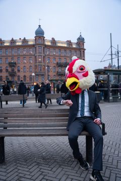 En person i kostym sitter på en bänk och bär ett stort kycklinghuvud som mask.