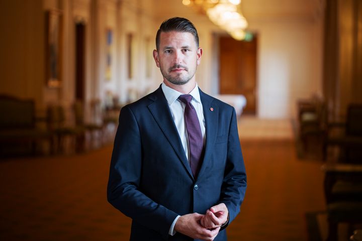 Samuel står och tittar in i kameran. Han har en blå kavaj och slips på sig. Han står i en riksdagskorridor.