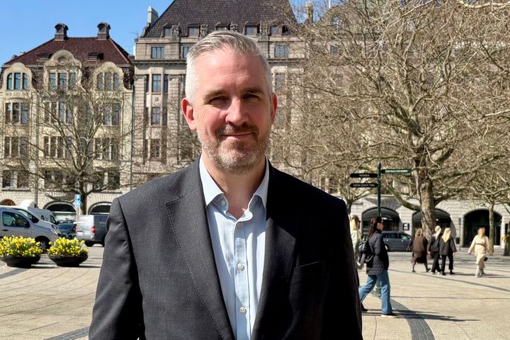 Bild på en man i kostym som står utomhus framför stora byggnader och träd.