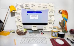 Beskrivning av ett skrivbord med en datorskärm täckt av post-it-lappar. Ett tangentbord, en nalle, en mugg och en papegoja syns också på bilden.