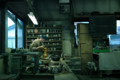 En man sitter i en studio och drejar. Framför sig arbetar han med en gul vas och längs väggen står rader av vaser och krukor i lera.